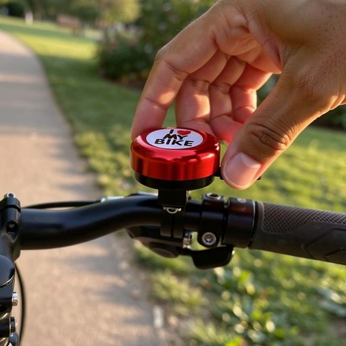 Eine Hand klingelt mit einer roten Fahrradklingel mit der Aufschrift „I Bike“. Weg und Gras im Hintergrund.