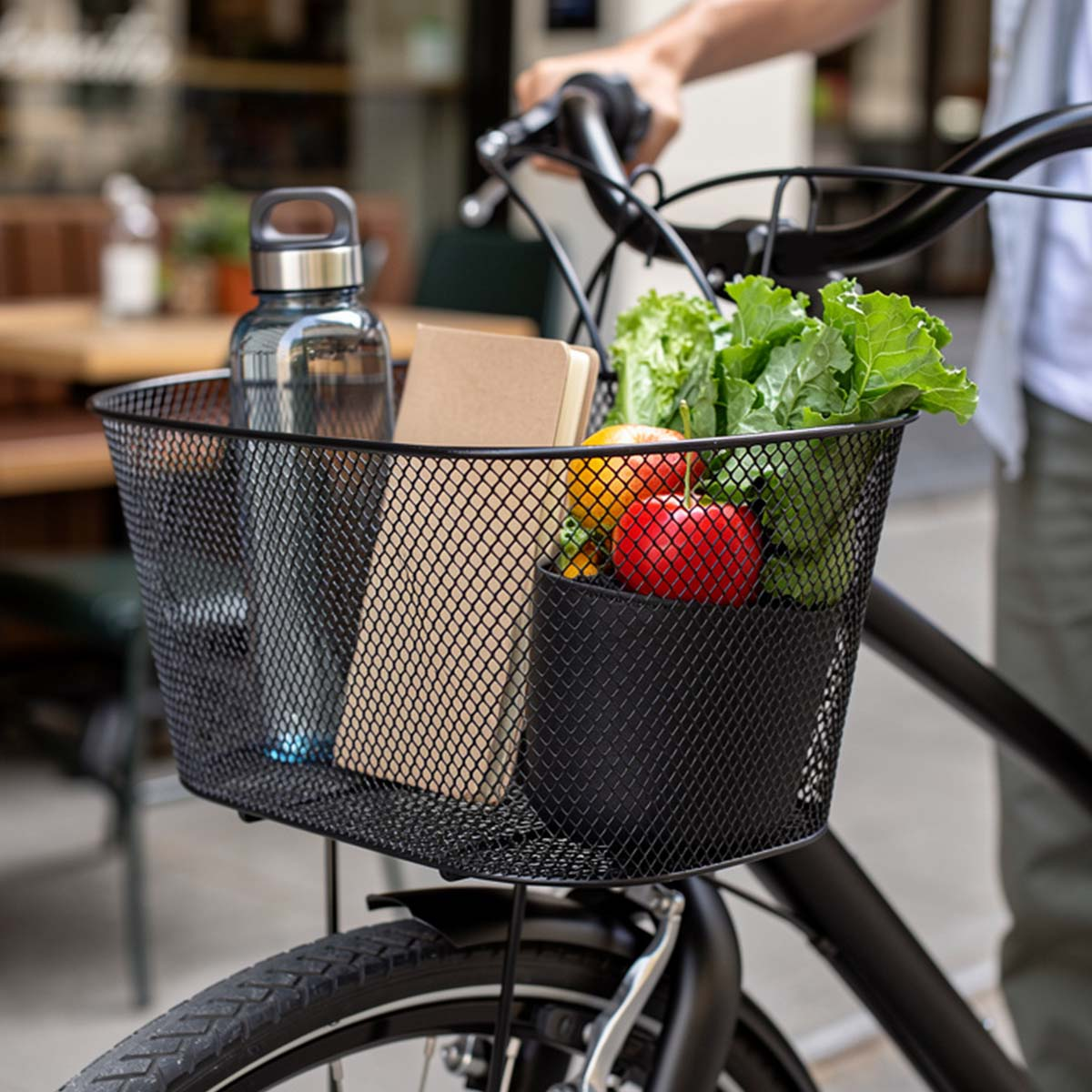 Fahrradkorb mit Wasserflasche, Buch und Lebensmitteln, von einer Person gehalten.