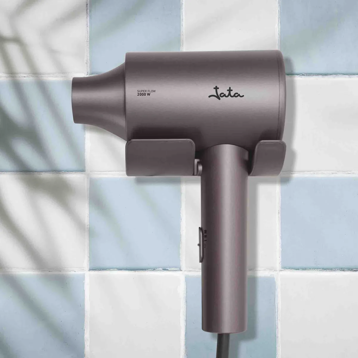 Un secador de pelo gris con el logo 'Jata' cuelga de una pared de azulejos azules y blancos.