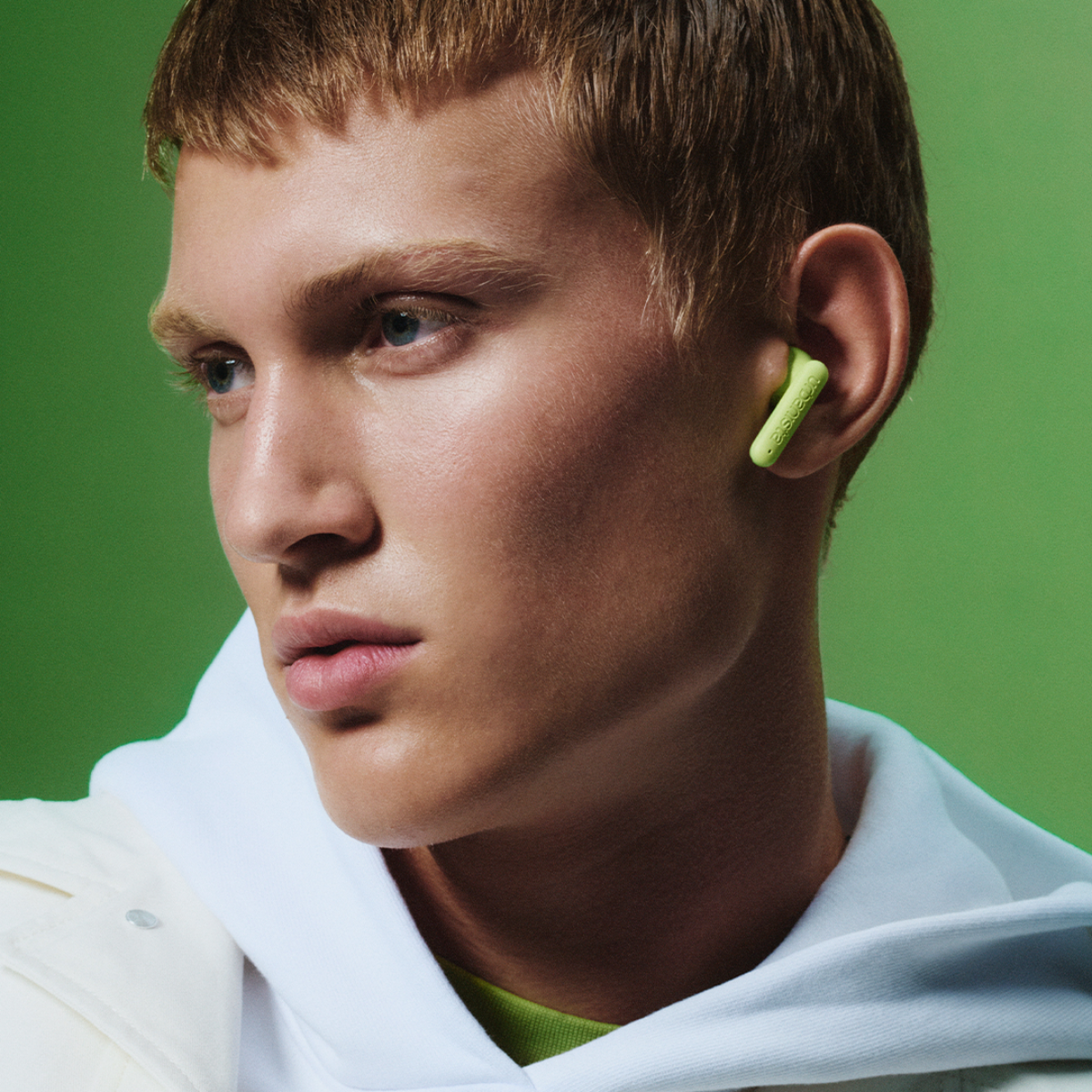 A man with short hair wears a white hooded jacket and green earbuds.