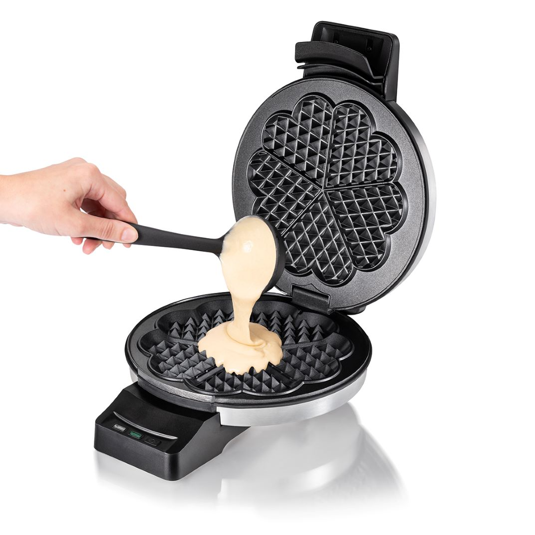 Una mano versa pastella con un mestolo in una piastra per waffle aperta a forma di cuore.