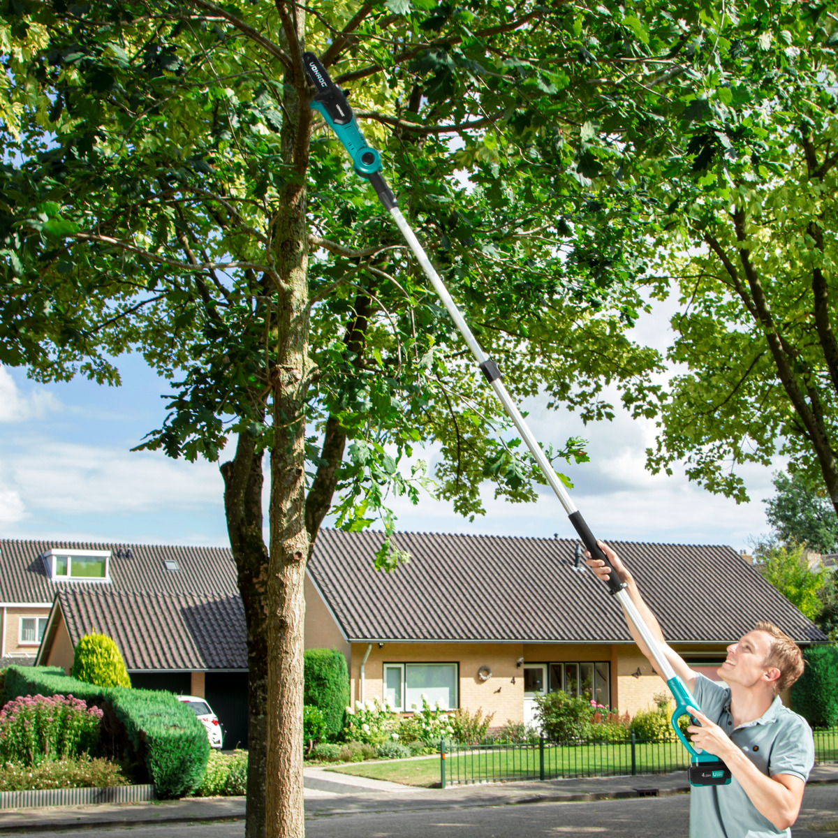 Man trims branches with a pole saw in a suburban neighborhood with houses.
