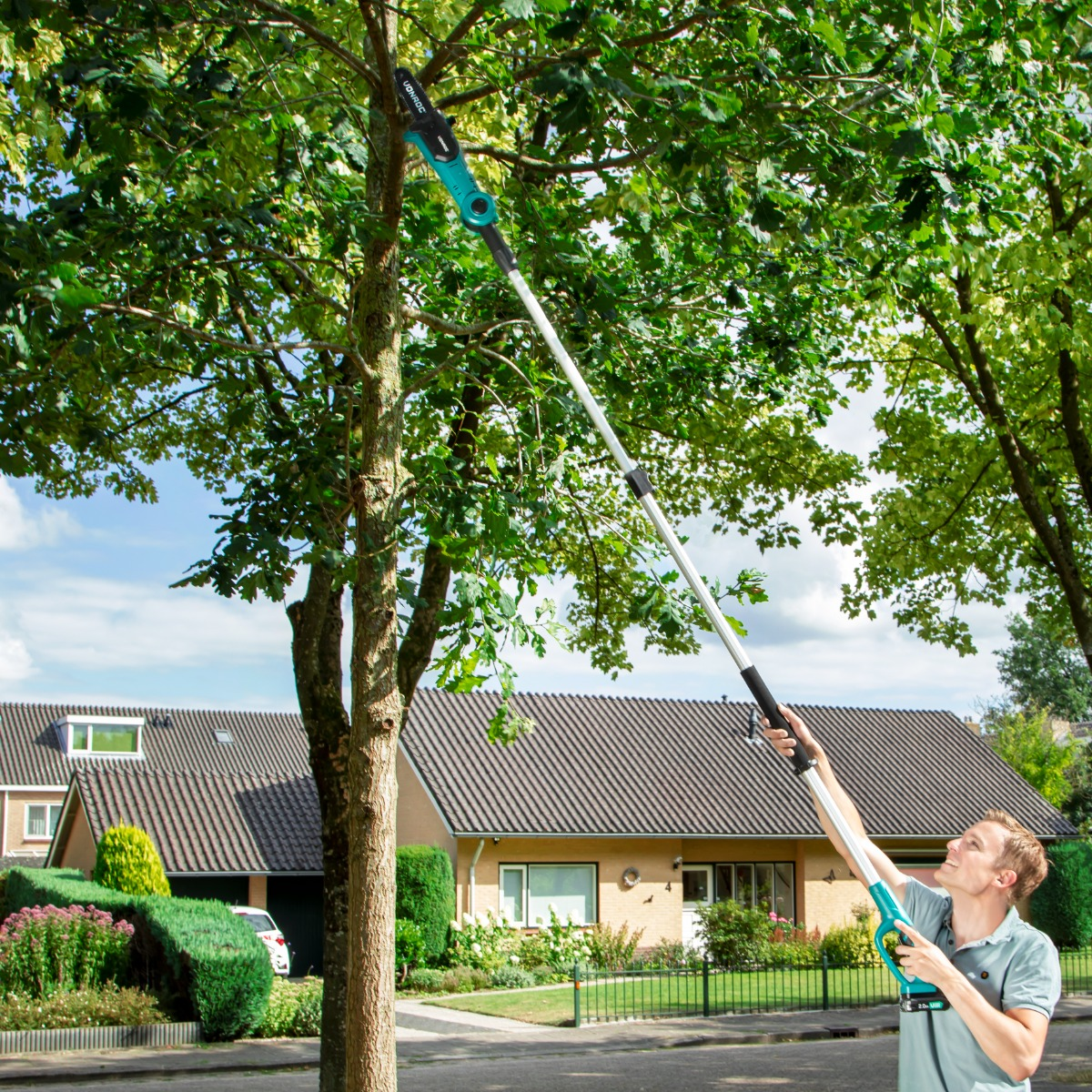 Man with pole saw cuts tree in suburban neighborhood.