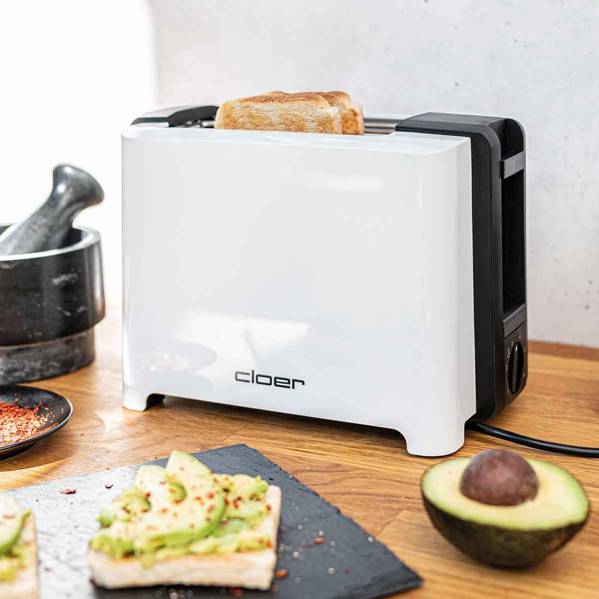 Biały toster z dwiema kromkami tostów, obok awokado i przypraw.