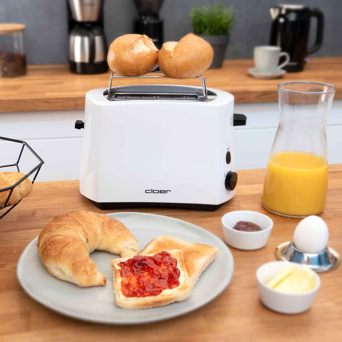 Ein weißer Toaster mit Brötchen, daneben ein Croissant, Toast mit Marmelade, Saft und Ei.