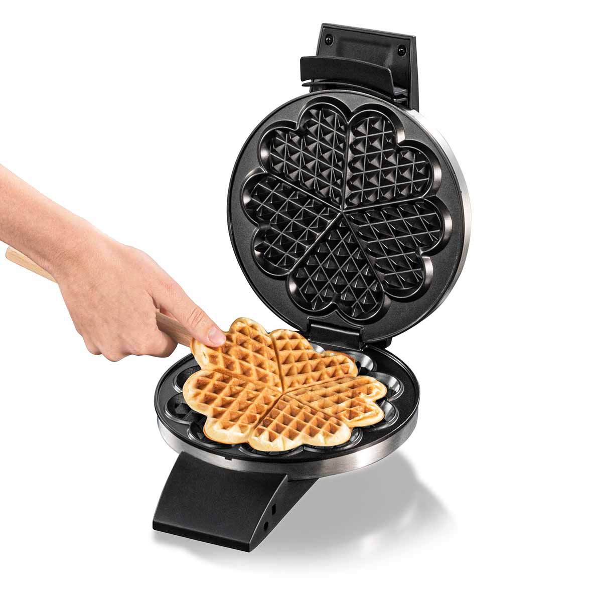 Ręka używa drewnianego narzędzia do wyjmowania gofra w kształcie serca z otwartej gofrownicy.