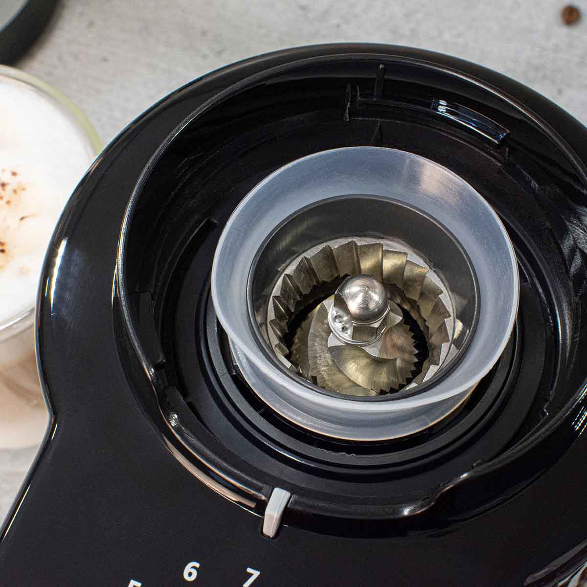Nahaufnahme des Mahlwerks einer Kaffeemühle mit einer Tasse Kaffee im Hintergrund.