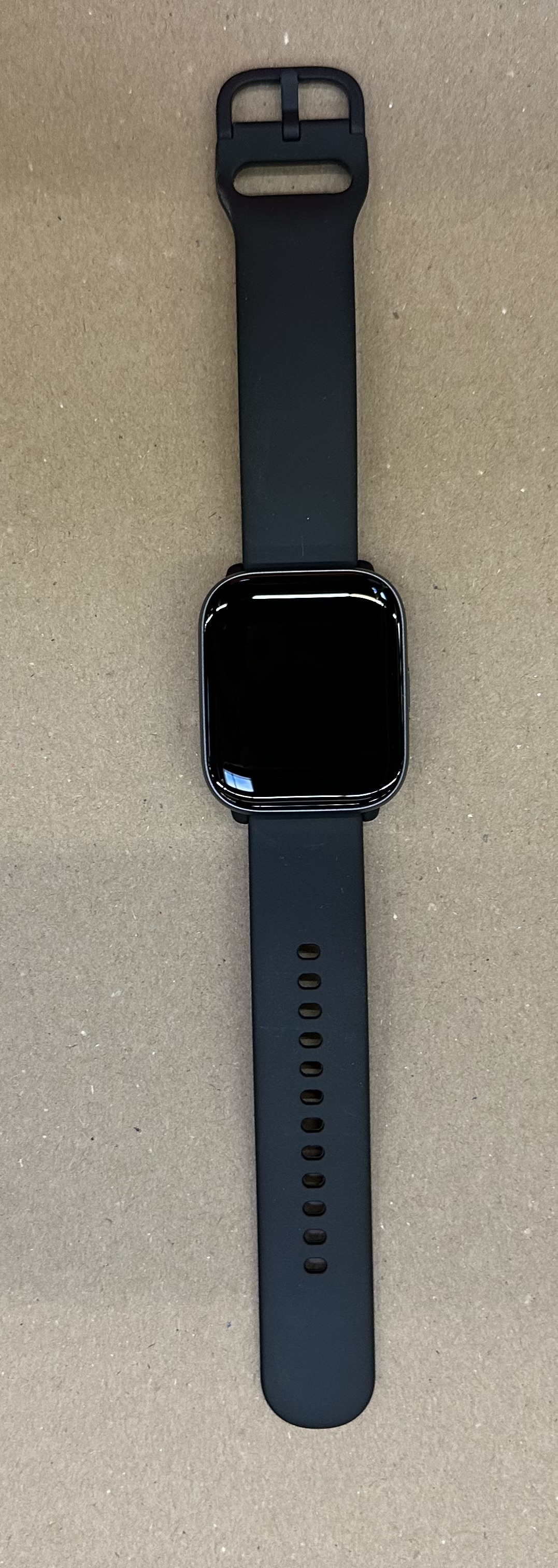 Uno smartwatch nero con schermo rettangolare e cinturino giace su una superficie marrone.