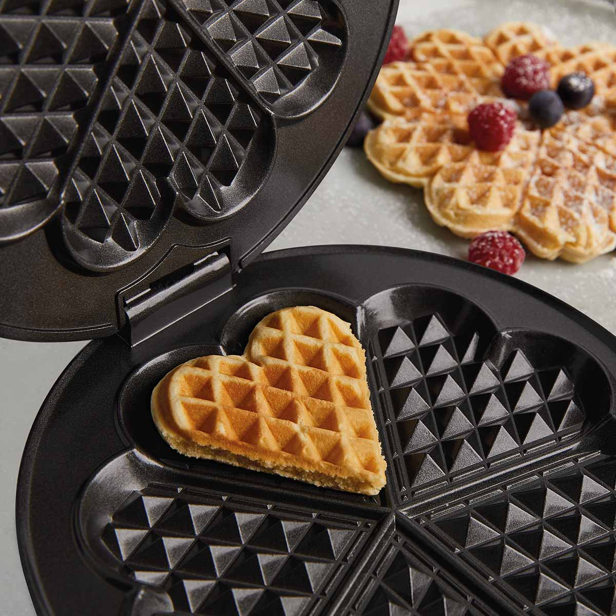 Gofrownica w kształcie serca z jednym gofrem w środku i więcej gofrów z jagodami w tle.