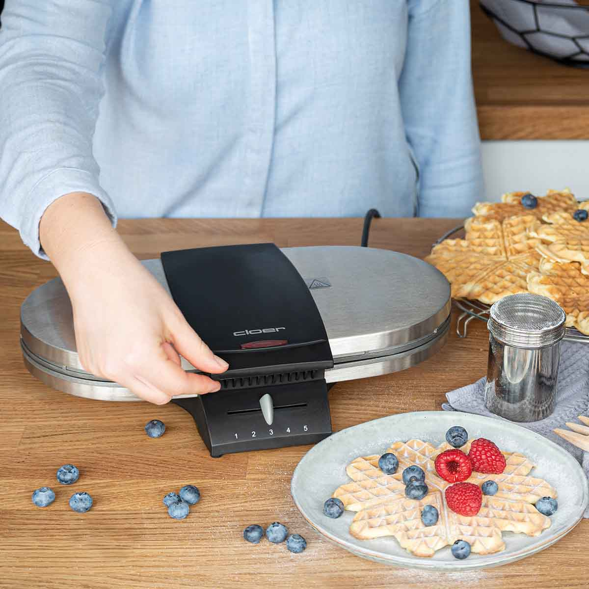 Dłoń reguluje stopień zrumienienia gofrownicy. W pobliżu znajdują się jagody i gofry.