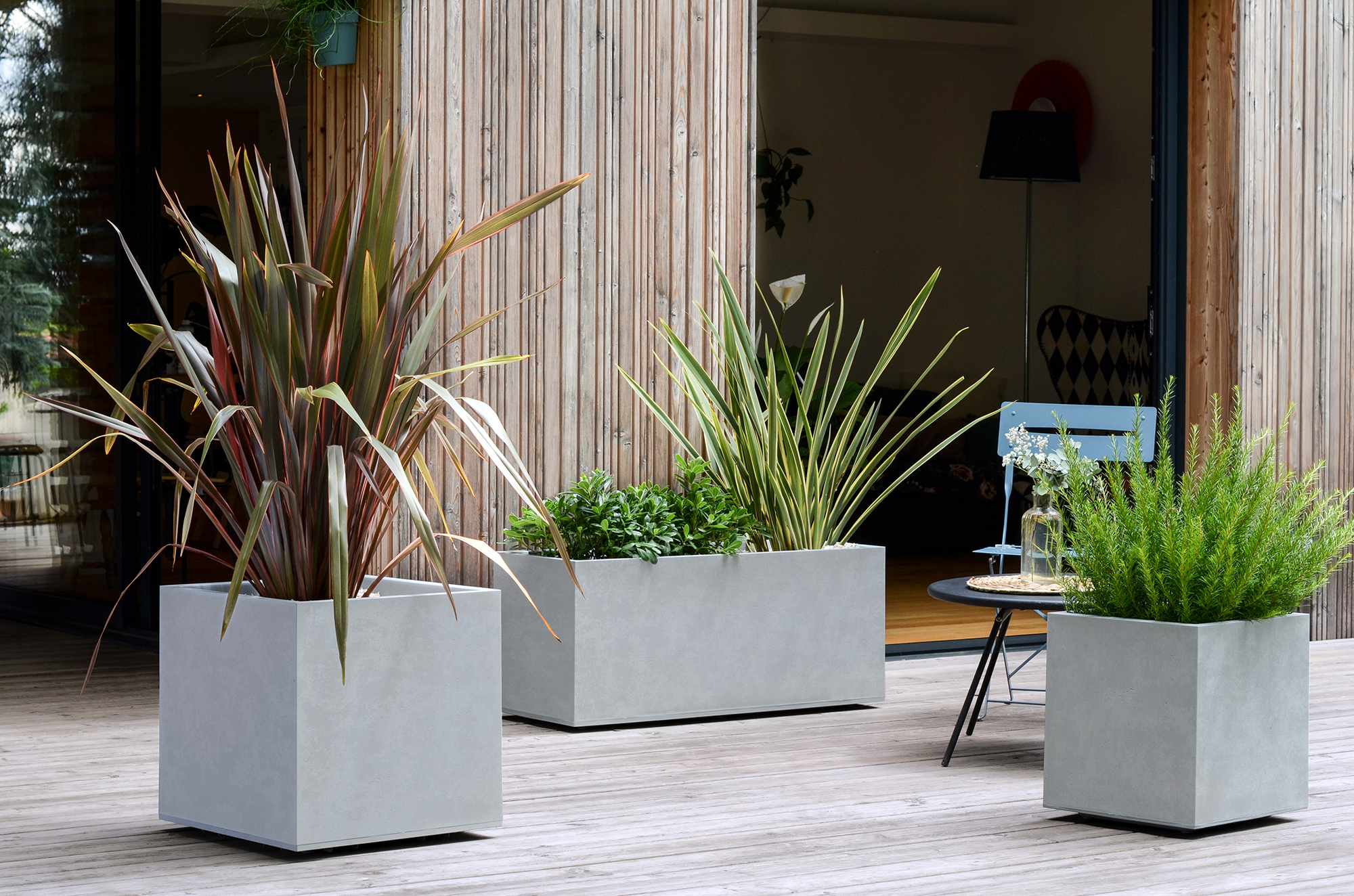 Moderne Betonpflanzkübel mit Pflanzen auf einer Holzterrasse vor einem Holznebengebäude.