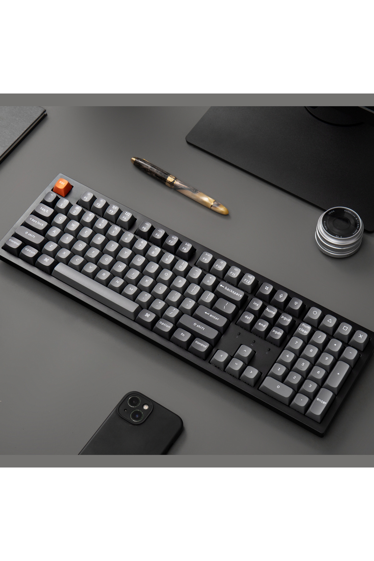 Czarna klawiatura z pomarańczowym klawiszem ESC, długopisem, telefonem i obiektywem.