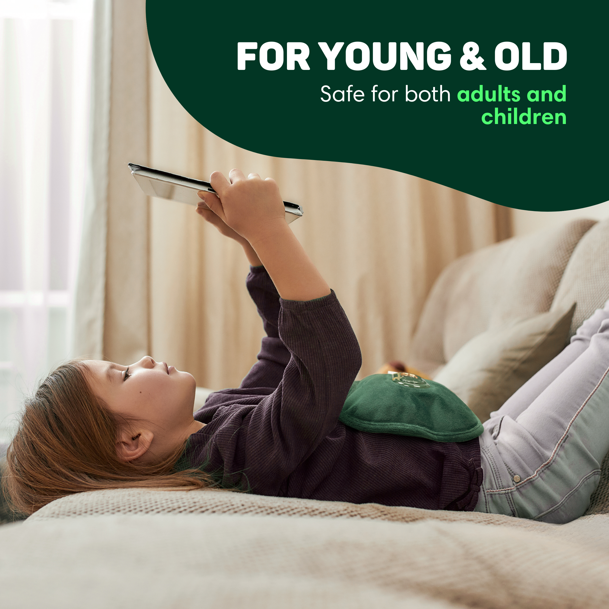 A child lies on a sofa holding a tablet. A green hot water bottle is on their stomach.