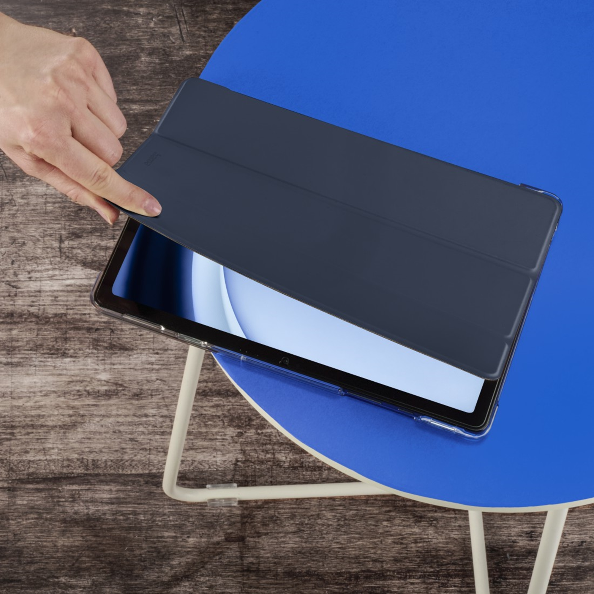Ręka zamyka ciemnoniebieskie etui na tablet na niebieskim stole.