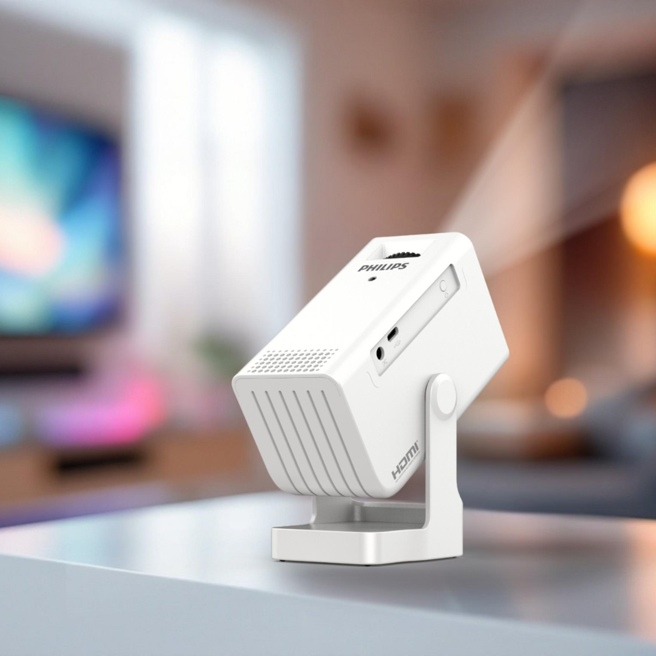 Biały projektor Philips stoi na stole. W tle niewyraźny telewizor.
