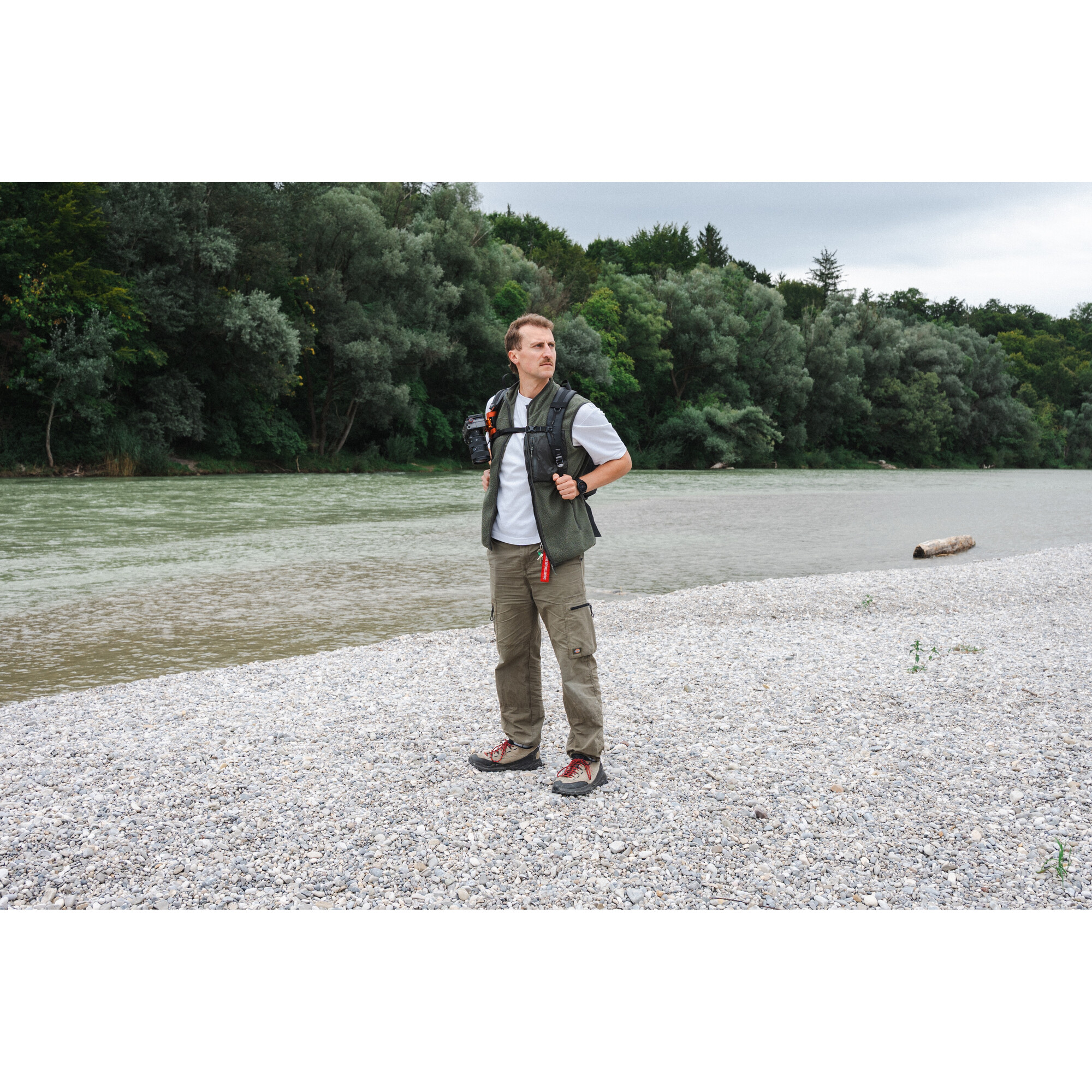 Ein Mann mit Weste und Rucksack steht am Ufer eines Flusses.