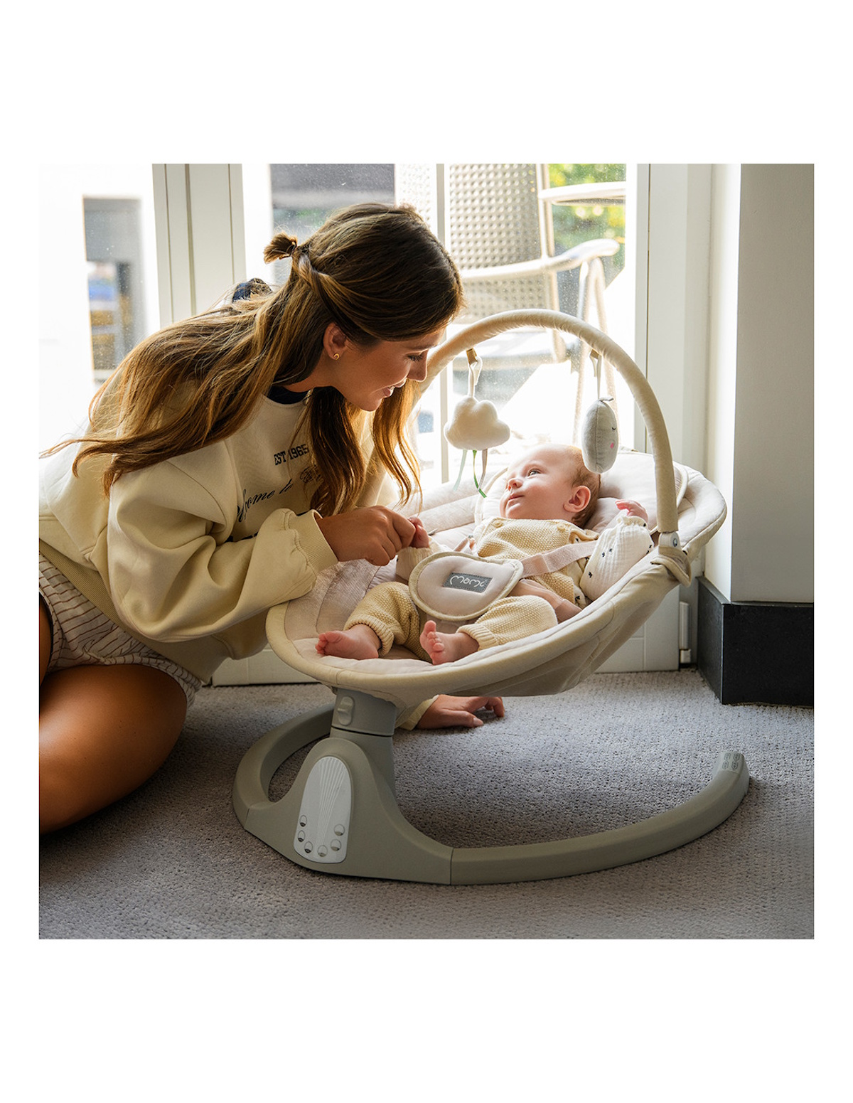 Eine Frau kniet vor einem Baby in einer Babyschaukel, die mit Spielzeug behängt ist.