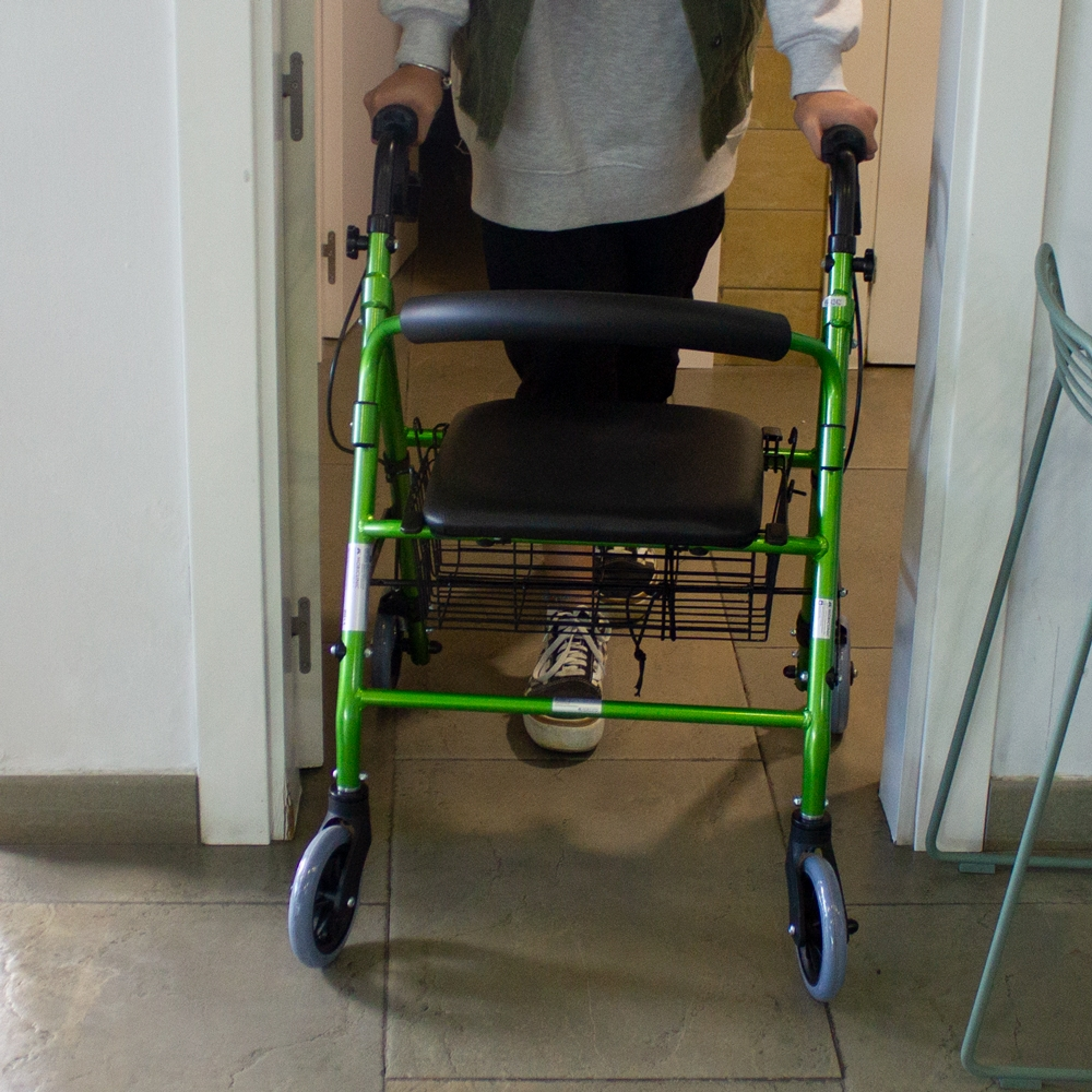 Andador verde con asiento y cesta, sostenido por una persona con zapatillas.