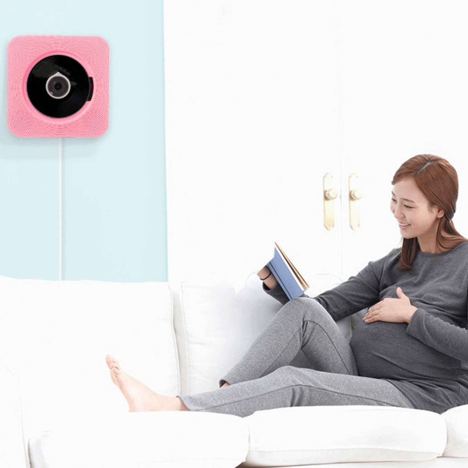 Eine schwangere Frau liest auf einem Sofa, ein rosa Babyfon hängt an der Wand.