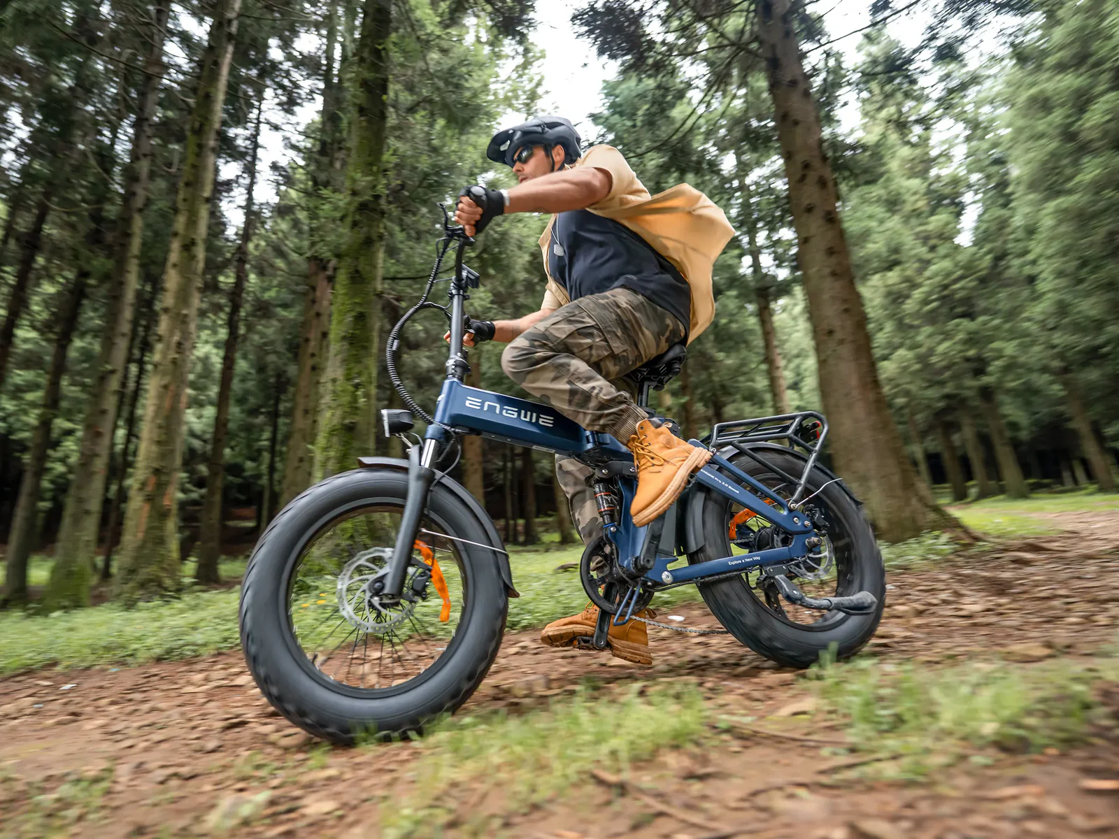Hombre montando bicicleta eléctrica azul en camino de bosque. Viste pantalones camuflados y botas.