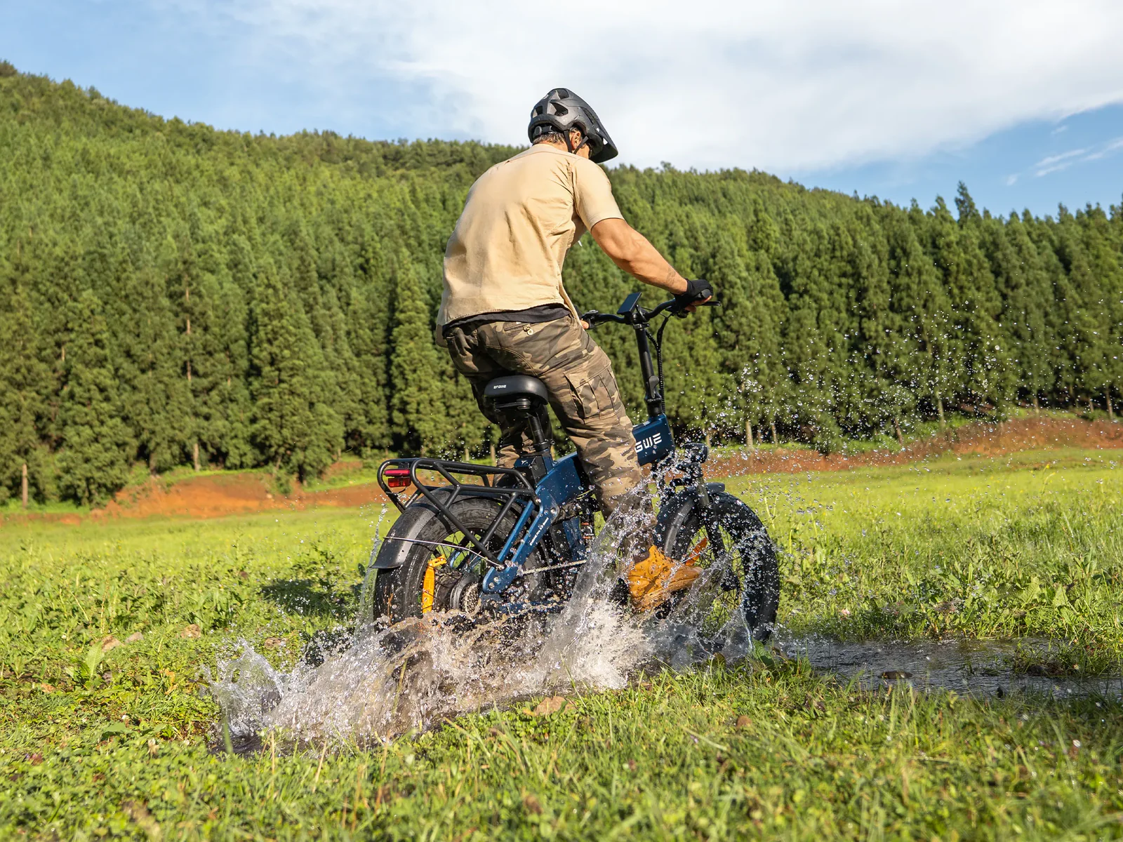 Uomo guida e-bike blu attraverso acqua in campo, alberi sullo sfondo.
