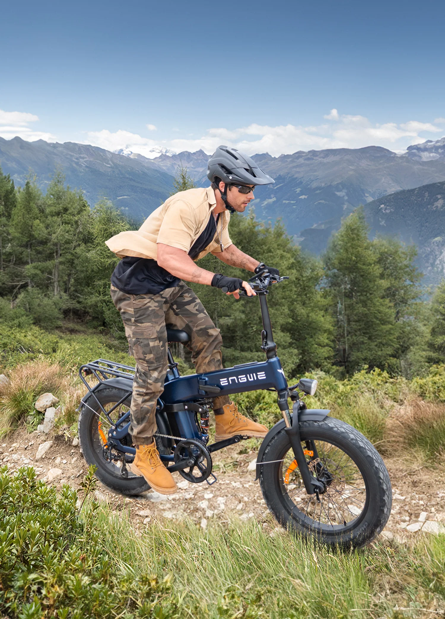 Uomo guida e-bike blu su sentiero montano con alberi e cime innevate.