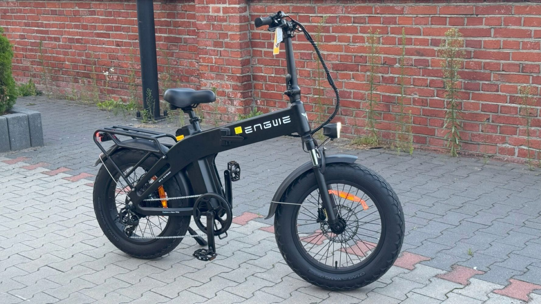 Una bicicleta eléctrica negra está aparcada en un camino pavimentado frente a una pared de ladrillos.