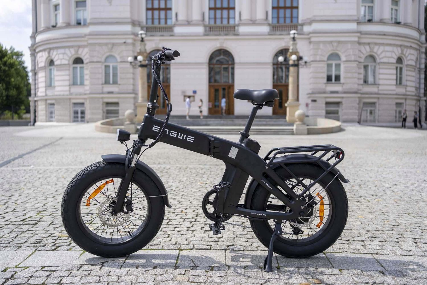 Una e-bike nera con pneumatici larghi si trova in una piazza acciottolata davanti a un grande edificio.