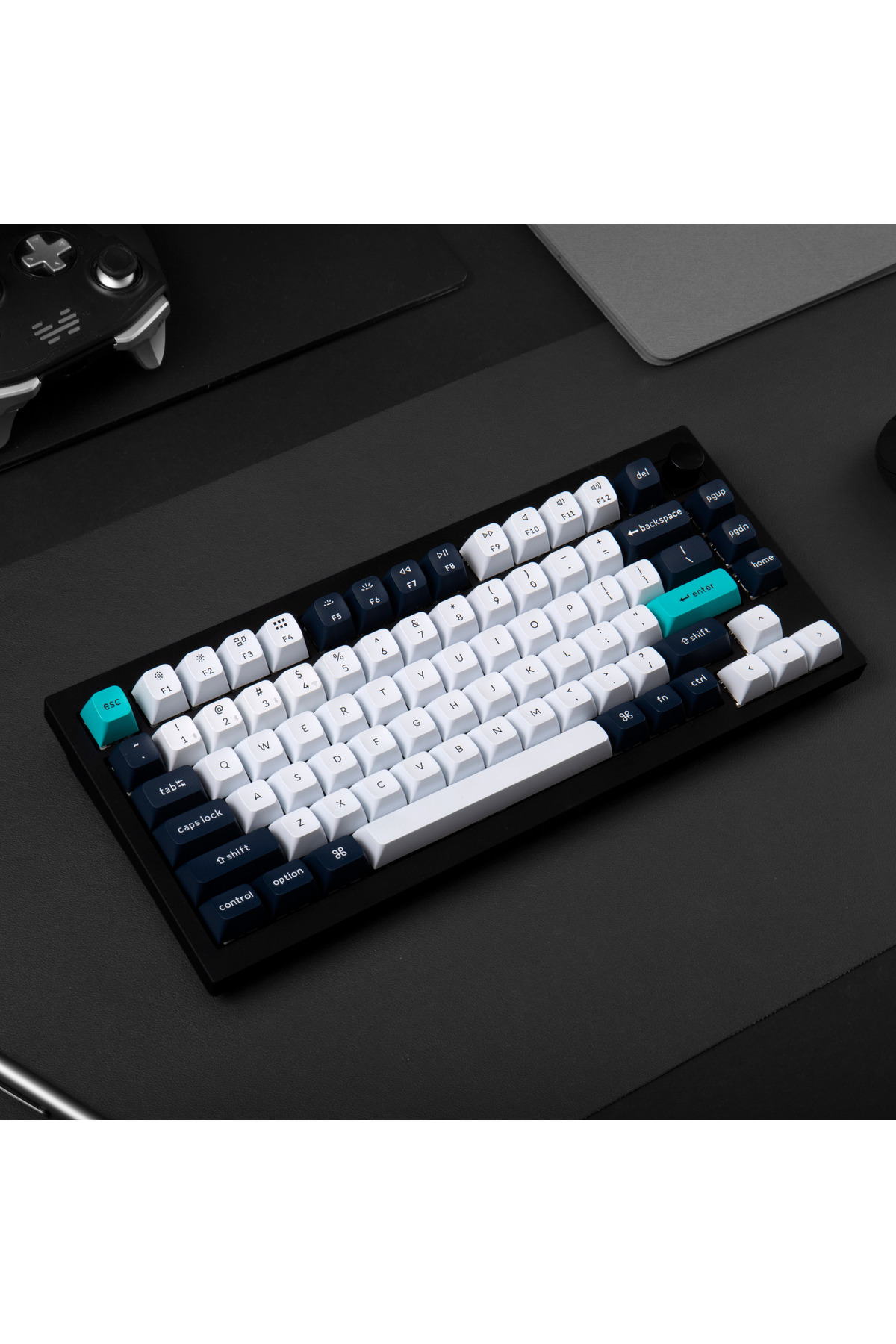 Un teclado mecánico negro con teclas blancas y azul oscuro sobre una alfombra negra. Un mando de juegos está cerca.