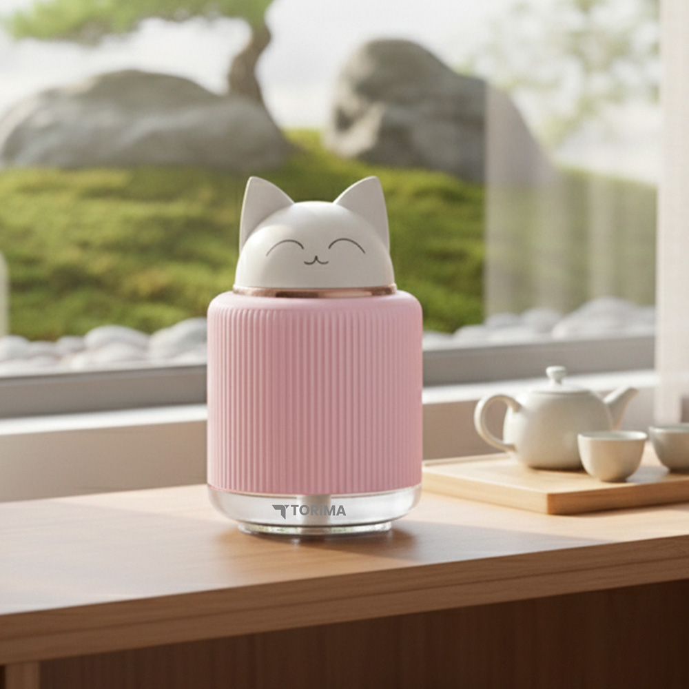Pembe kedi şeklinde bir difüzör, yanında çaydanlık ve fincanlarla ahşap bir masada duruyor.
