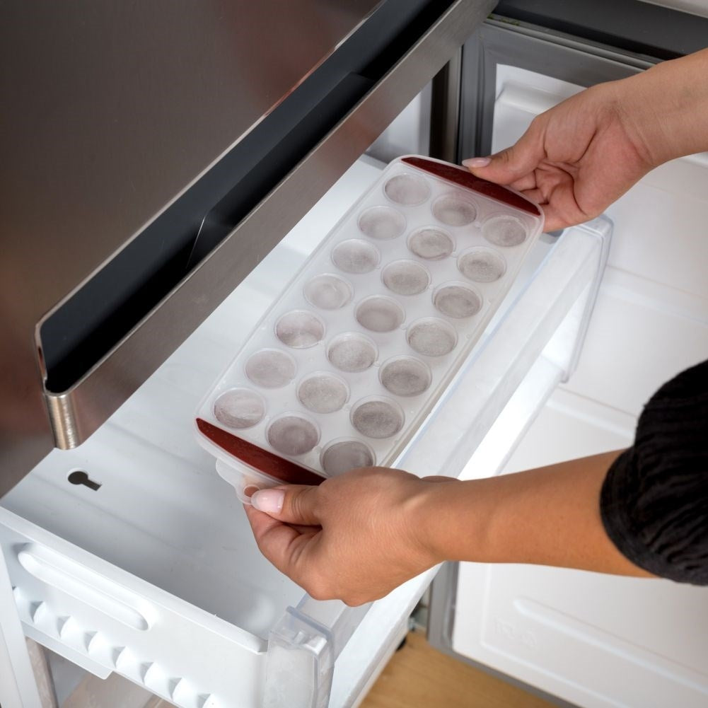 Manos colocando una cubitera en un refrigerador. La cubitera tiene muchos compartimentos redondos.