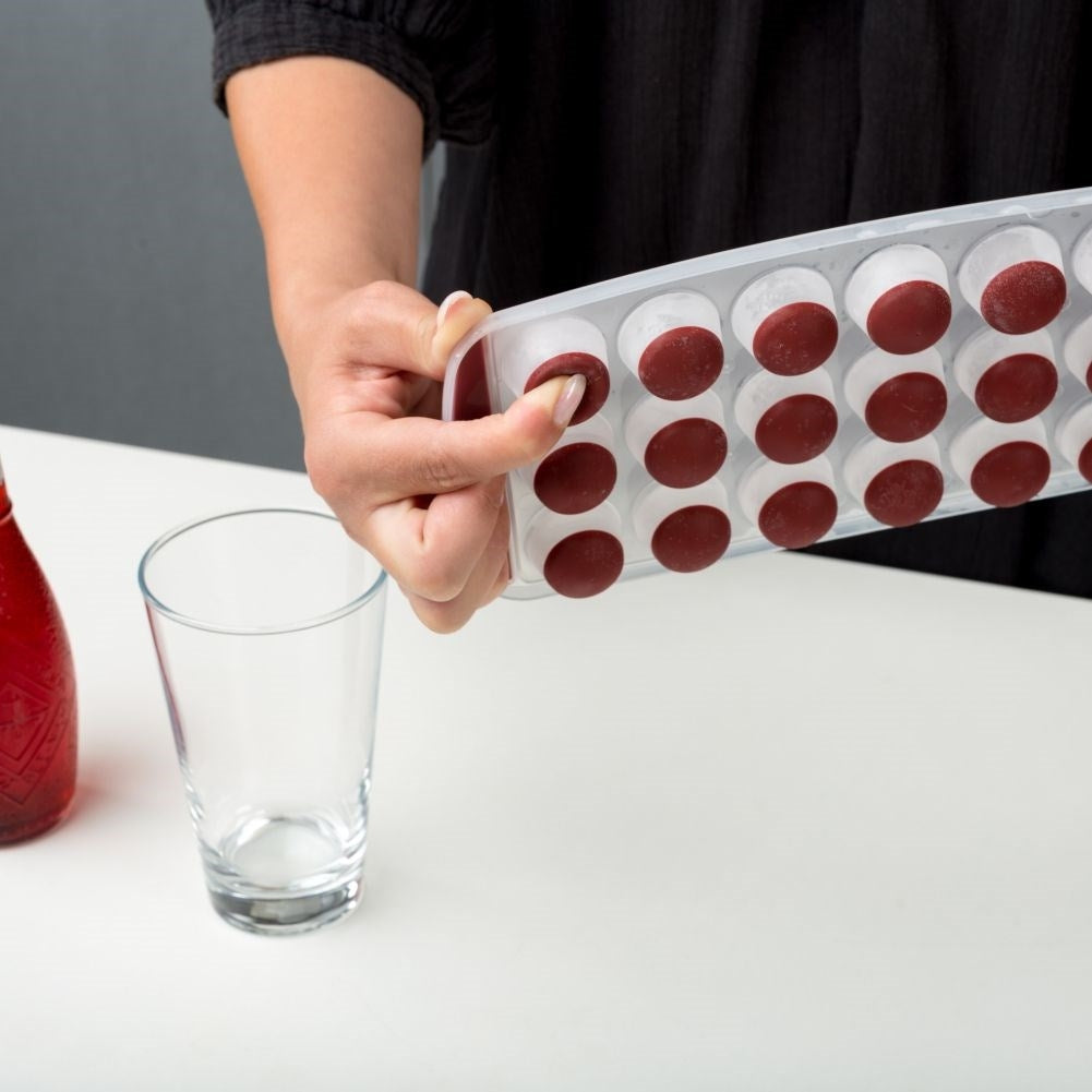 Mano sosteniendo molde para cubitos de hielo con cubos rojos y blancos sobre un vaso.