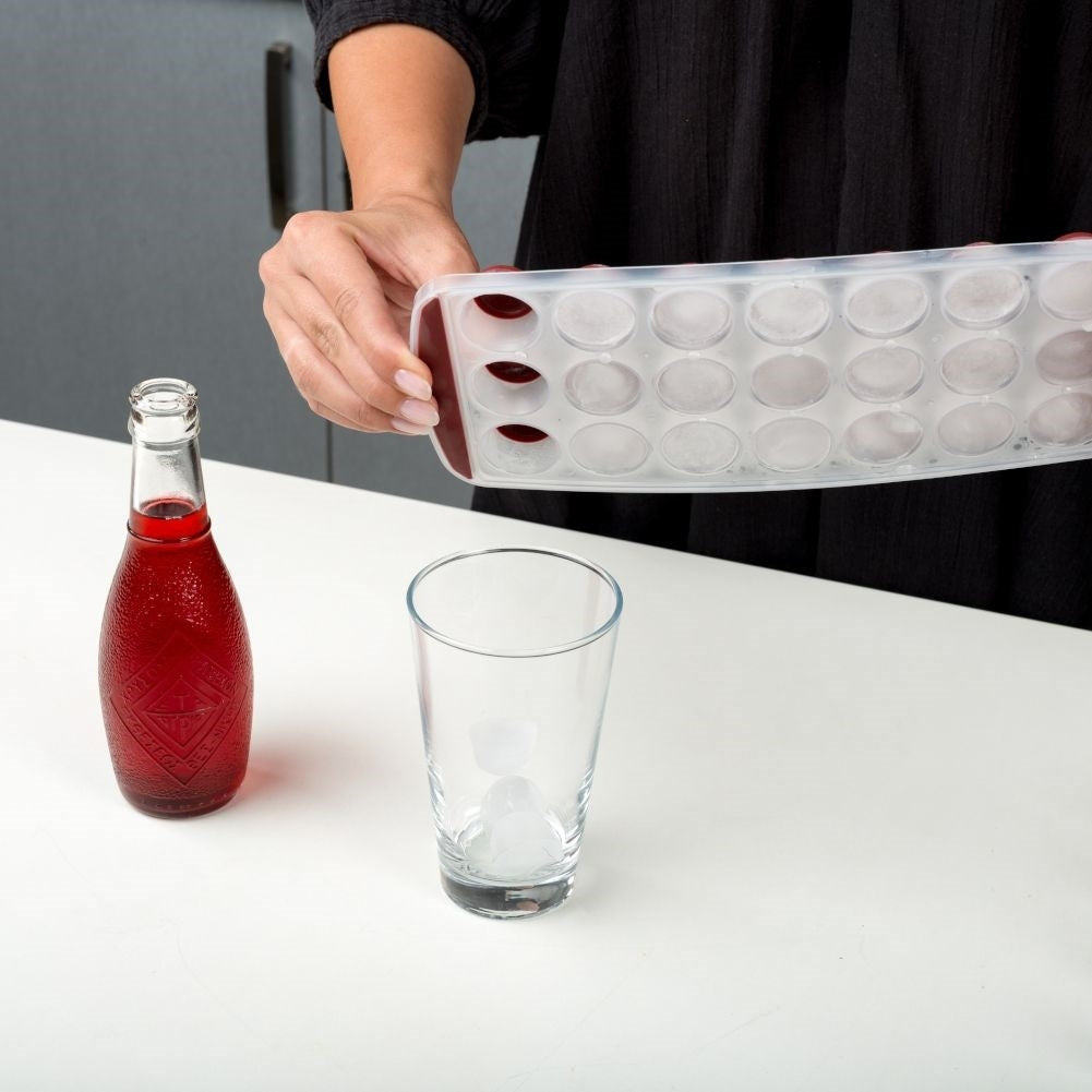 Una persona vierte cubitos de hielo de una bandeja en un vaso junto a una botella de refresco roja.