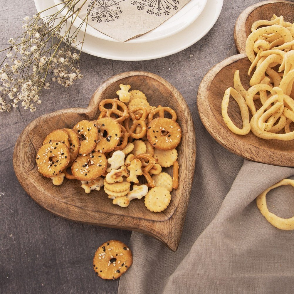 Bol de madera en forma de corazón con galletas variadas. Cerca hay otro bol con snacks en forma de anillo.