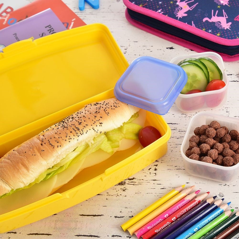 Una lonchera amarilla tiene un sándwich, con bocadillos y lápices de colores cerca.