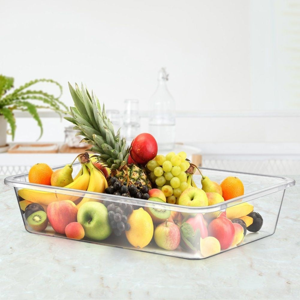 Un recipiente transparente lleno de frutas variadas: piña, manzanas, uvas, naranjas.