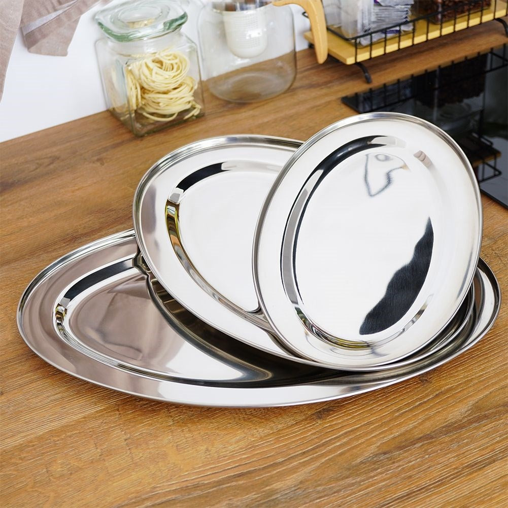 Tres platos ovalados de metal brillante sobre una mesa de madera. Al fondo, vasos y una estantería.