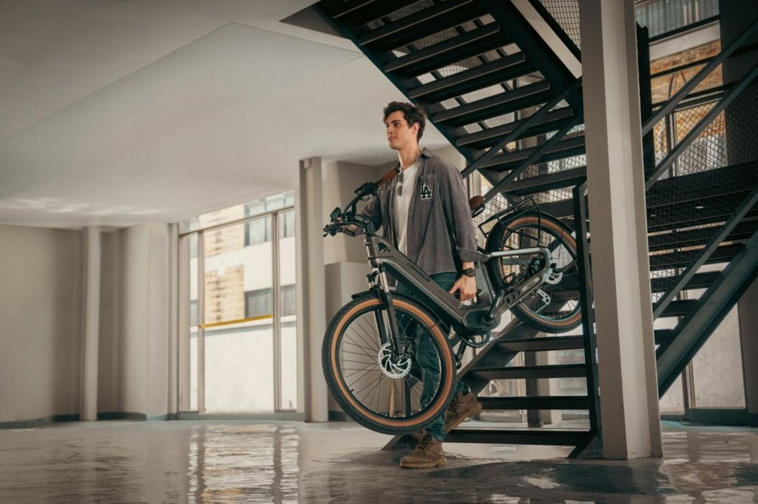 Un hombre sube una bicicleta gris por las escaleras. Lleva camisa y vaqueros.