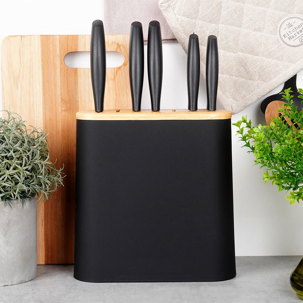 Un bloque de cuchillos negro con tapa de madera, junto a una tabla de cortar de madera y plantas.