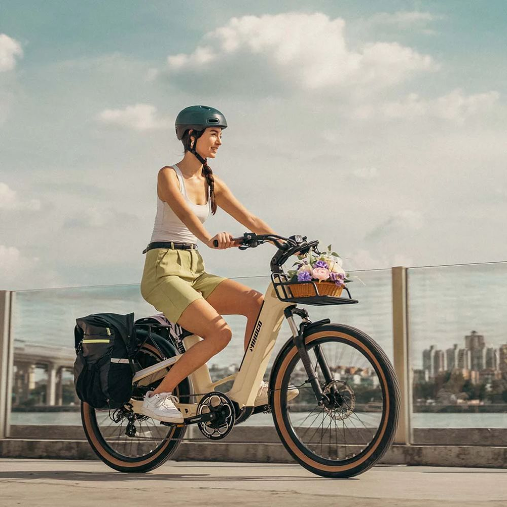 Mujer monta en bicicleta con cesta de flores, casco y alforjas, ciudad al fondo.