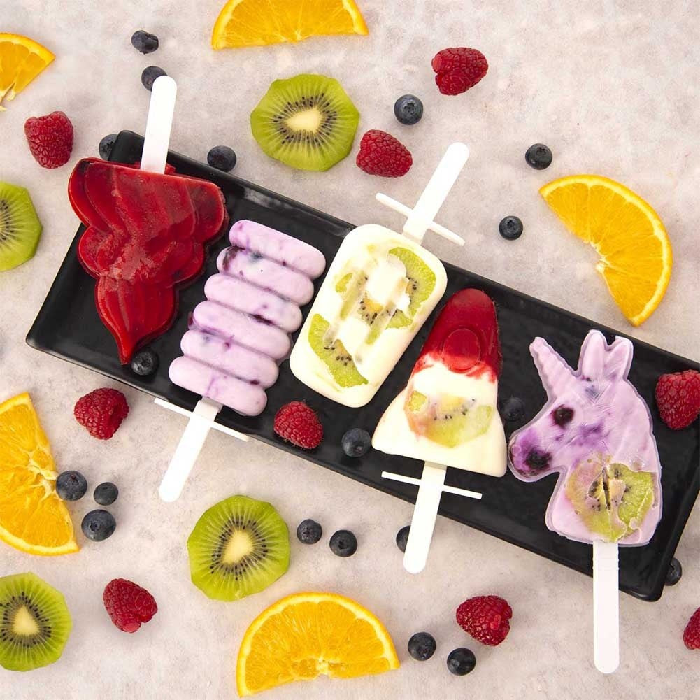 Cinco paletas coloridas con fruta en un plato negro, adornadas con bayas y naranjas.