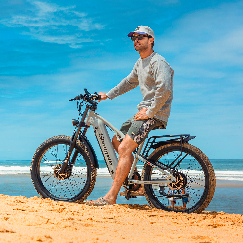 Mann auf einem E-Bike am Strand, mit Blick aufs Meer, bei blauem Himmel.