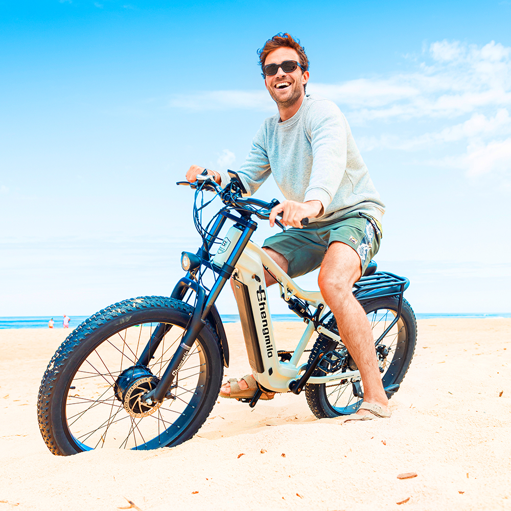 Ein Mann reitet ein E-Bike am Strand. Der Himmel ist blau mit Wolken.