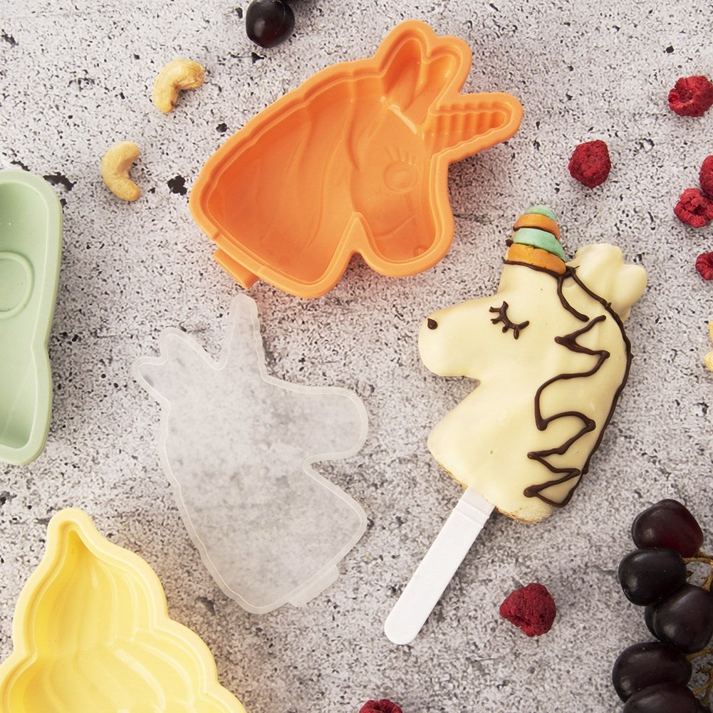Molde de helado de unicornio con helado terminado, moldes y fruta en superficie gris.