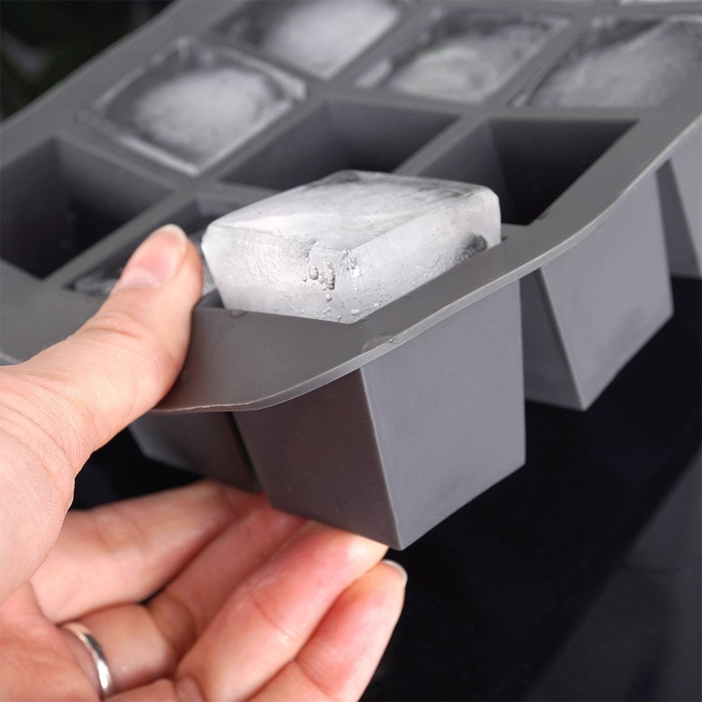Una mano saca un cubito de hielo de un molde de silicona gris con muchos cubos.