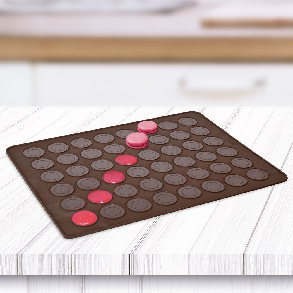 Alfombrilla de silicona marrón para macarons con macarons rosas en varias etapas de preparación sobre mesa de madera blanca.
