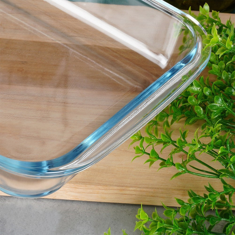 Una fuente de vidrio para hornear con borde azul sobre tabla de madera junto a plantas verdes.