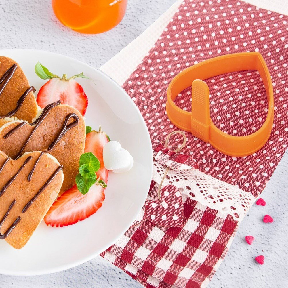 Tortitas en forma de corazón con fresas, chocolate y un cortador sobre un paño a cuadros.