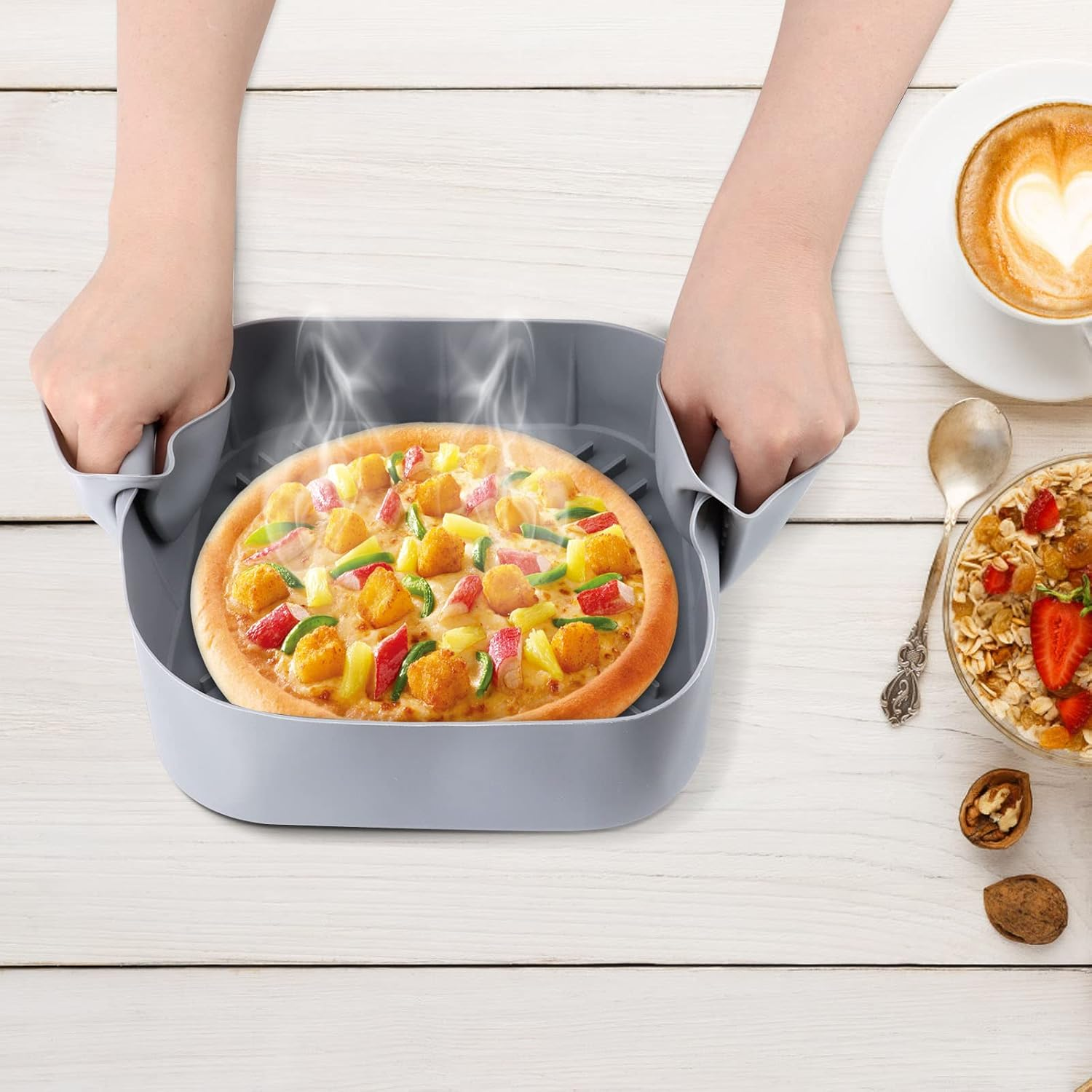 Hände halten eine graue Silikonform mit einer dampfenden Pizza und Frühstück auf dem Tisch.