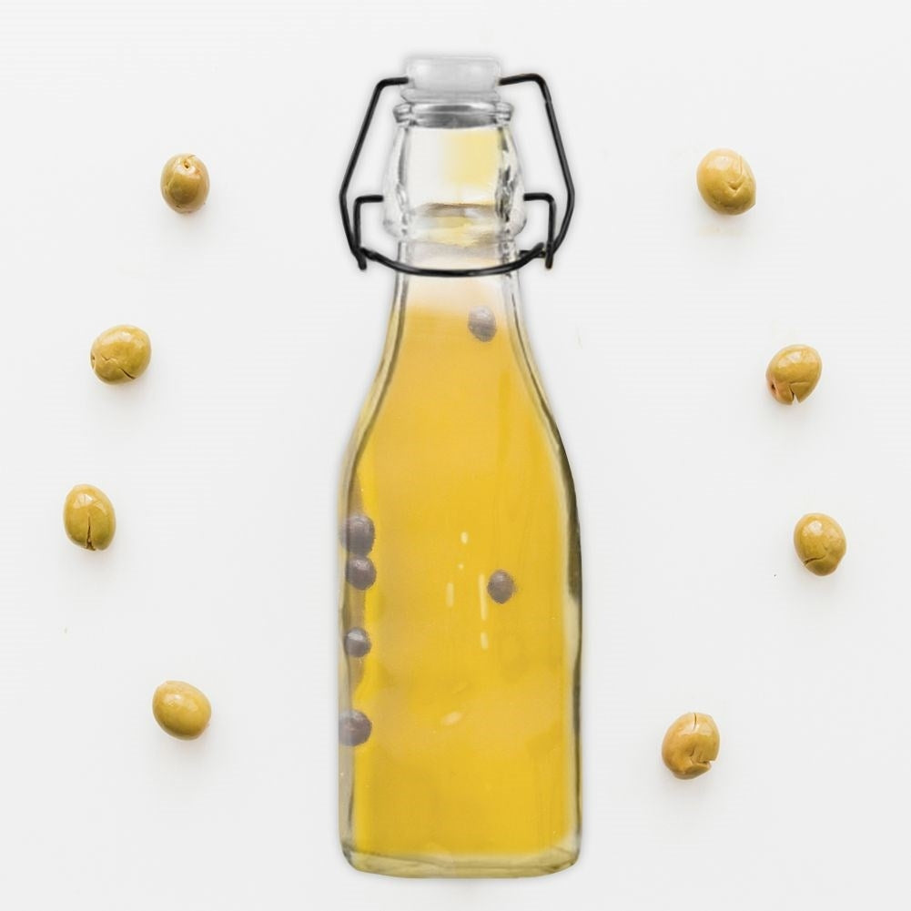 Una botella de vidrio con aceite amarillo y aceitunas dispuestas a su alrededor.